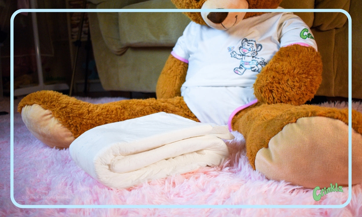 Image of folded BetterDry adult diaper in front of a large teddy bear wearing a Crinklz onesie.